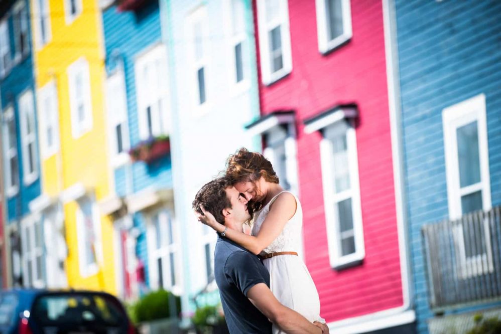 A man lifts his soon to be wife in the air as they have their photograph taken in front of colorful houses.