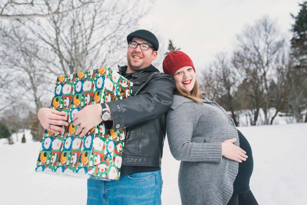 A couple pose happily in front of the lens, dad to be holding a big present and mommy to be holding her belly.