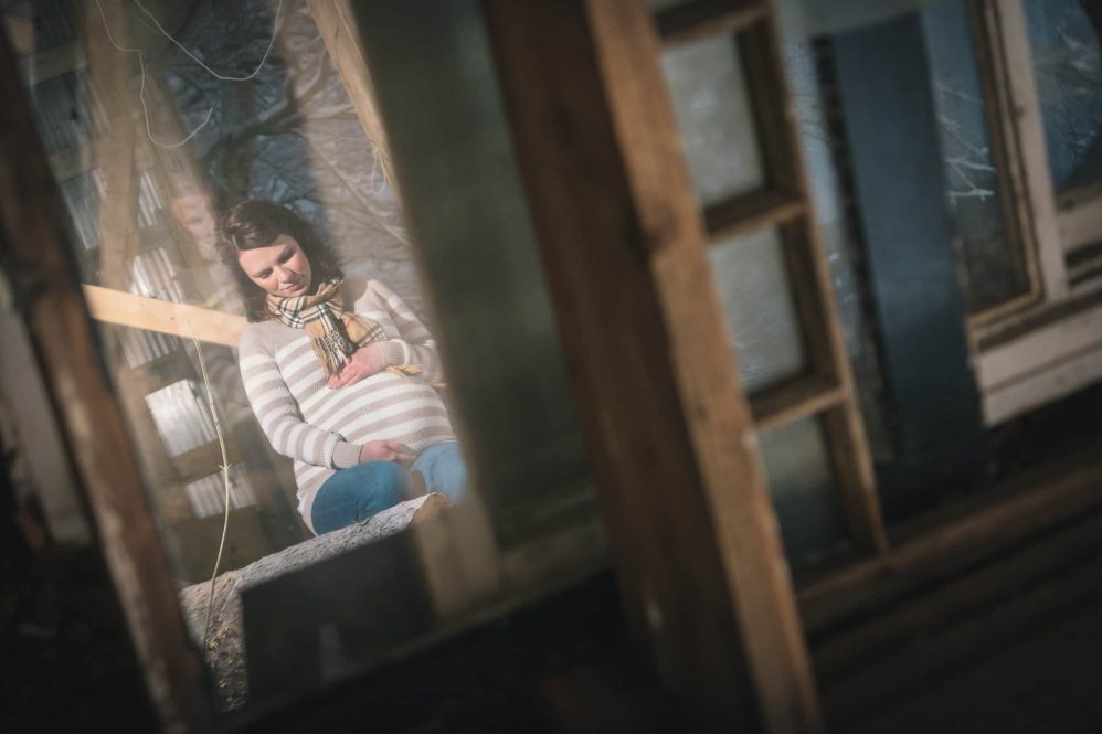 In this maternity photo a mother to be holds her stomach while the photo is shot in the reflection of an hold window.