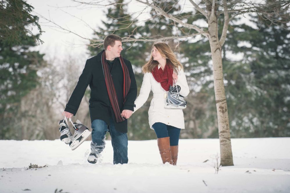 A newly engaged couple holding hands walks through the snow with their ice skates over their shoulder.