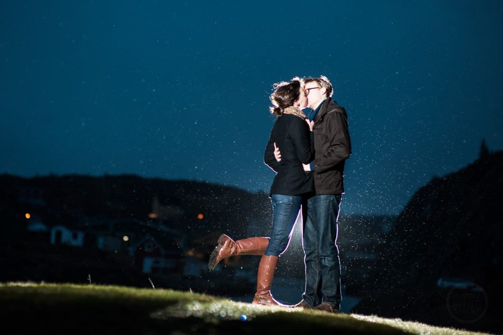 With raindrops backlit by a remote flash, a young couple poses for a photo atop a small hill.