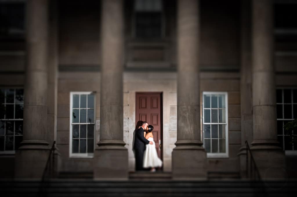 In a timeless fine art style photography a bride and groom kiss against a red door with a soft light illuminating the colomns of The Colonial Building.