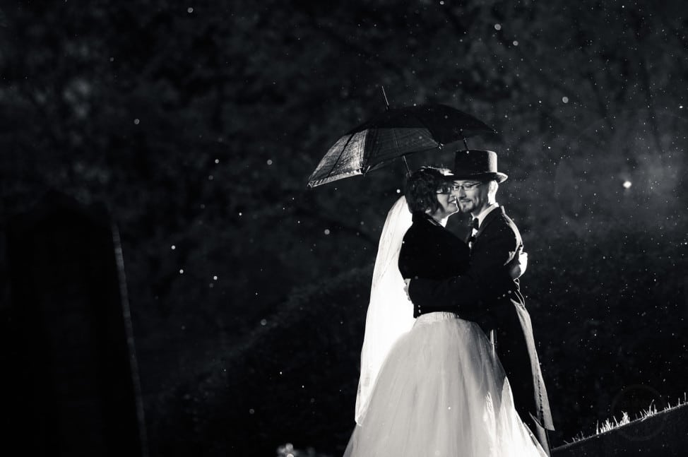 Inky blacks are delicately teased with backlit raindrops as bride and groom embrace under an umbrella.