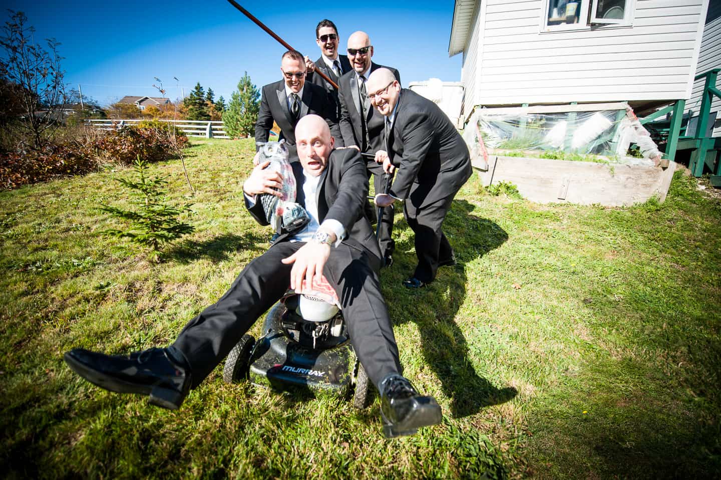 A group of groomsmen push the groom-to-be down a hill on a lawnmower while he holds an owl statue in this hilarious Newfoundland wedding photo.