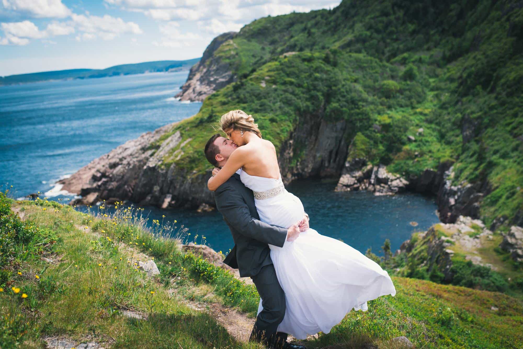 A bride jumps into her grooms arms to kiss him with the colorful coastline of Newfoundland and Labrador stretching off in the background
