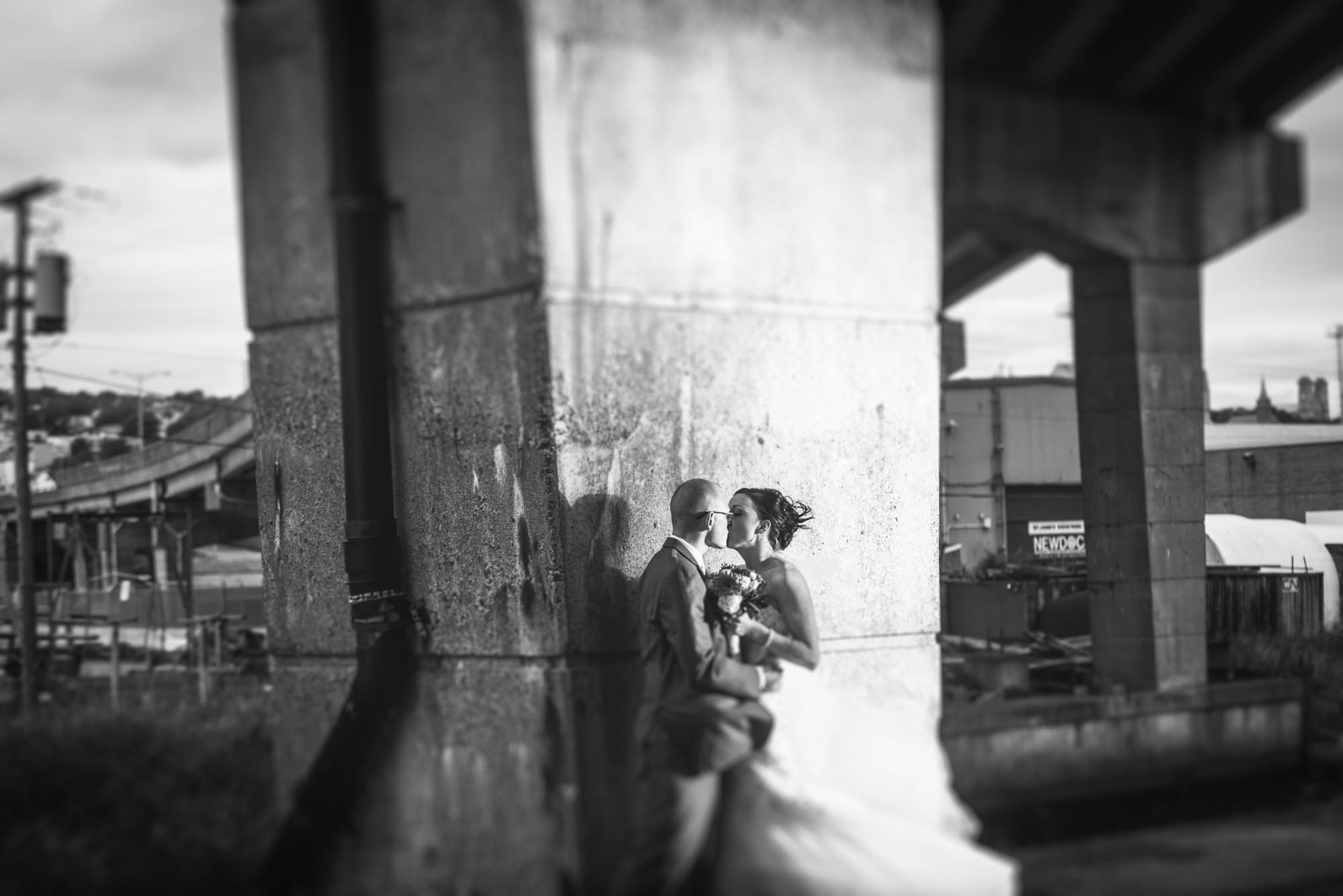 Under a overpass a bride and groom kiss in a moody and grundgy black and white photograph