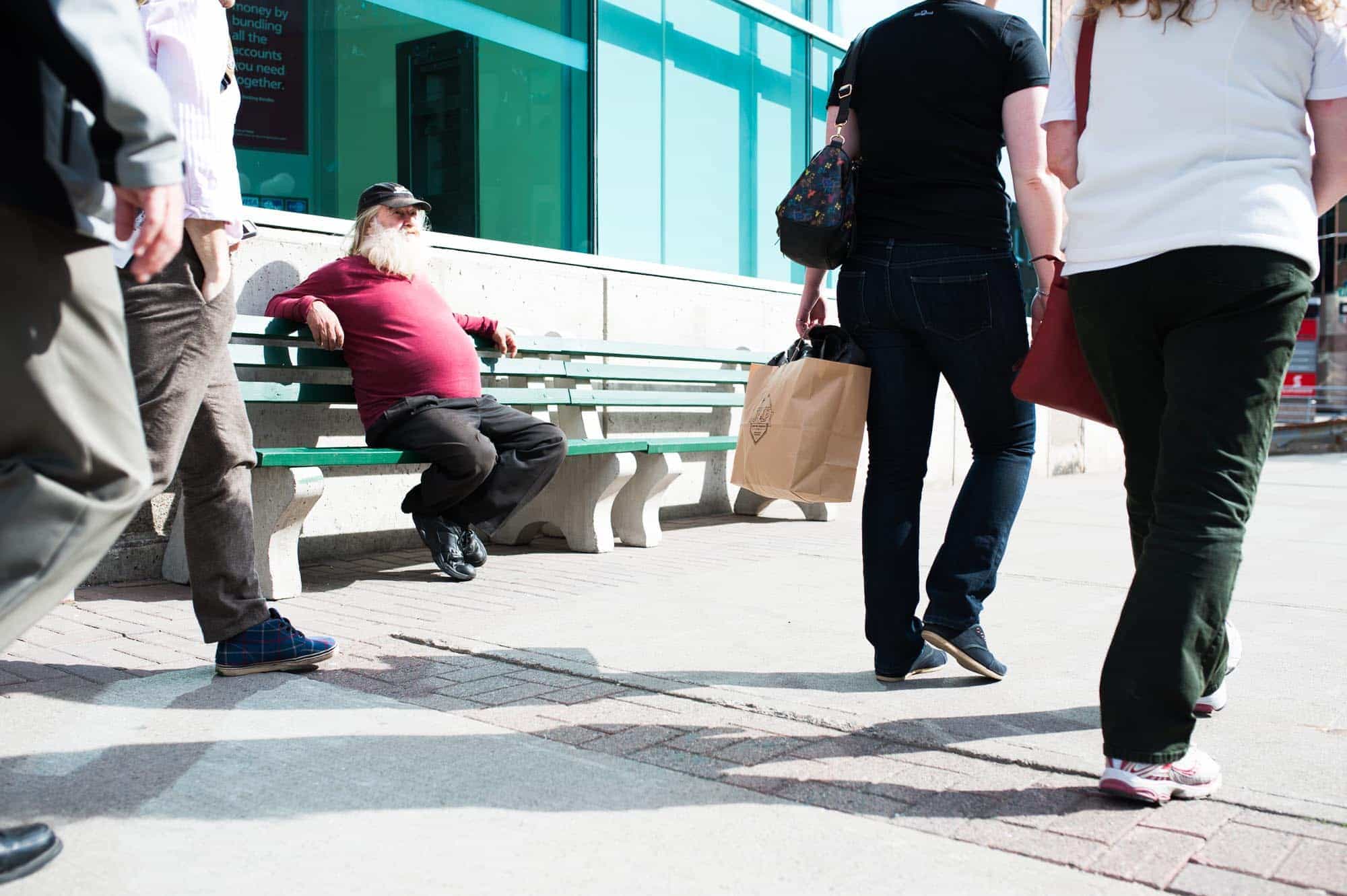 A photograph on Water Street in St. John's shows a large belly man in a red shirt and white beard relaxing on a park bench as people pass by.