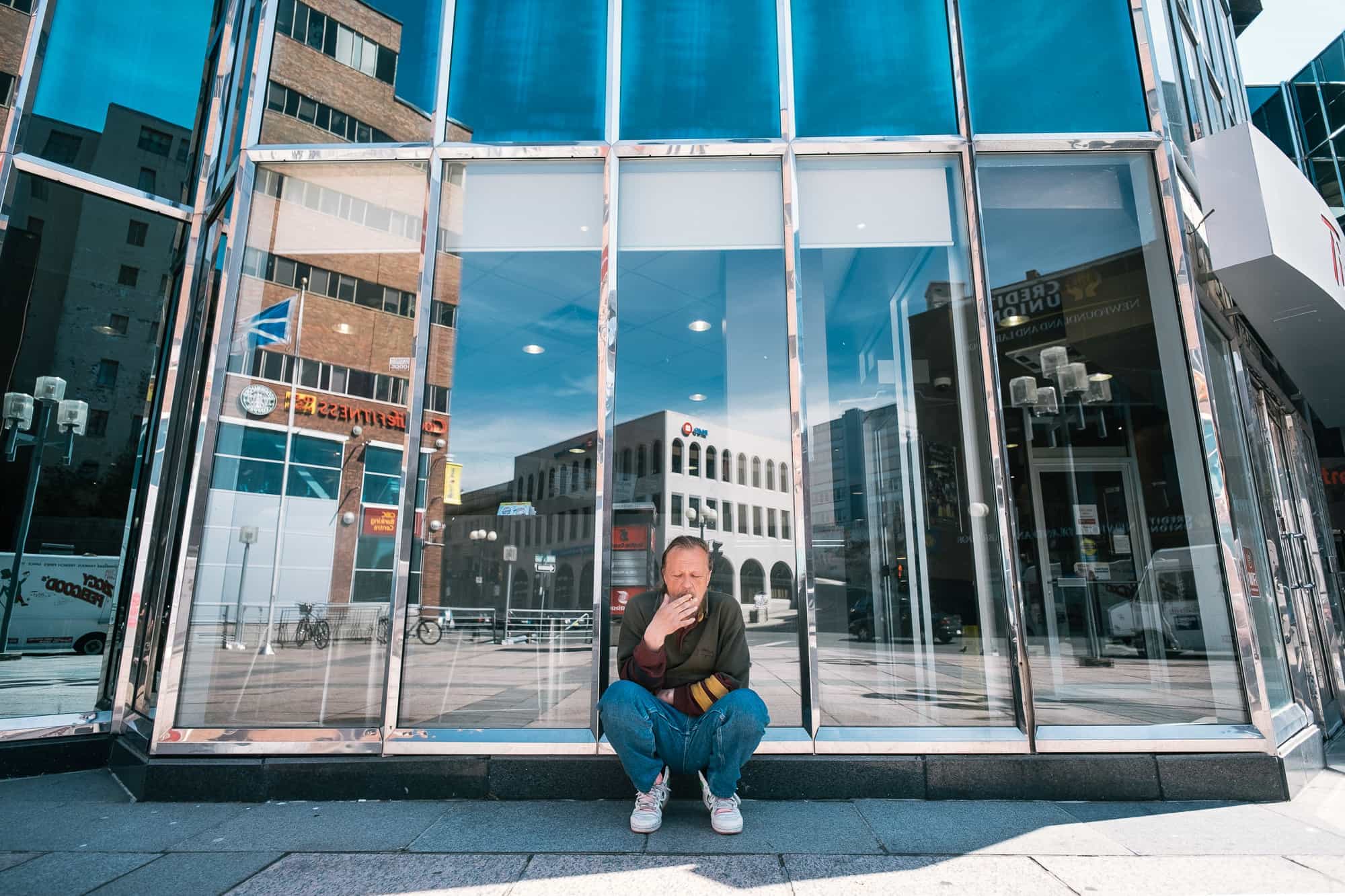 A man sits against a glass window smoking a cigarette for this St. John's street photograph.