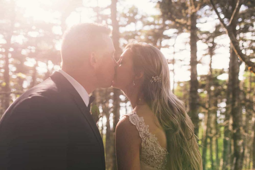 A bride kisses her groom in the forest as the sun peeks through.