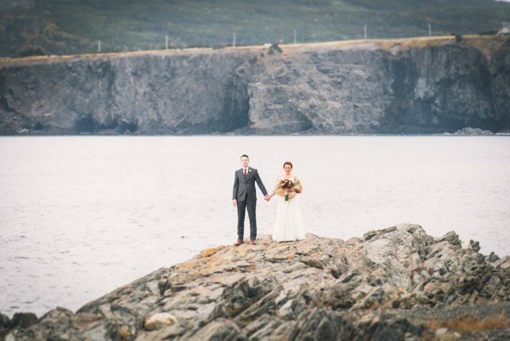 A bride and groom hold hands posing on the edge of the coastline just inches from the freezing atlantic ocean.