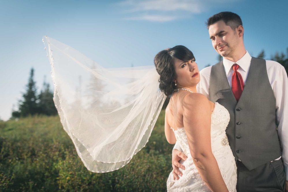 A bride's veil is caught in the wind as her groom looks happily upon her.